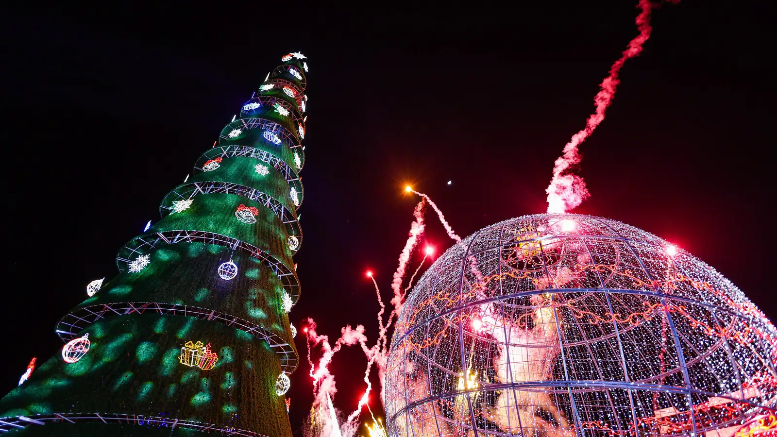 The giant Christmas tree and the giant ornament lit up at night, with fireworks rising between them