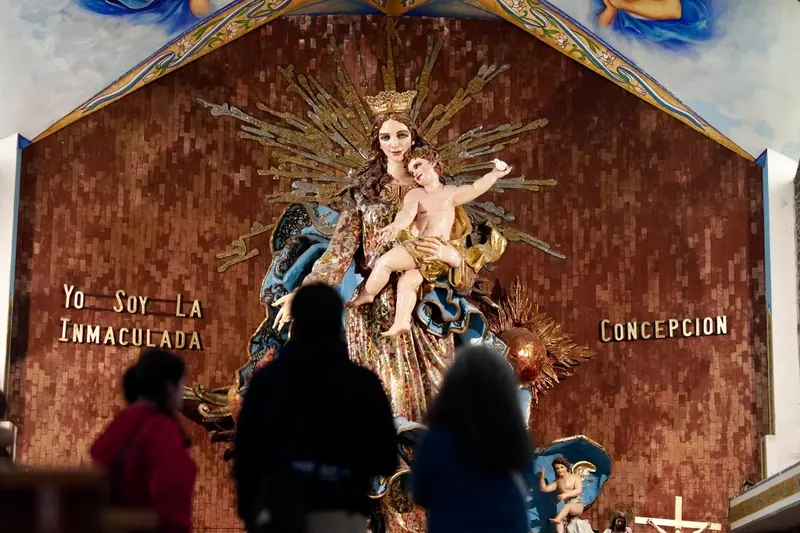 Visitantes de pie frente a la gran imagen de la Inmaculada Concepción; sobre el muro dorado se lee «Yo soy la Inmaculada Concepción».