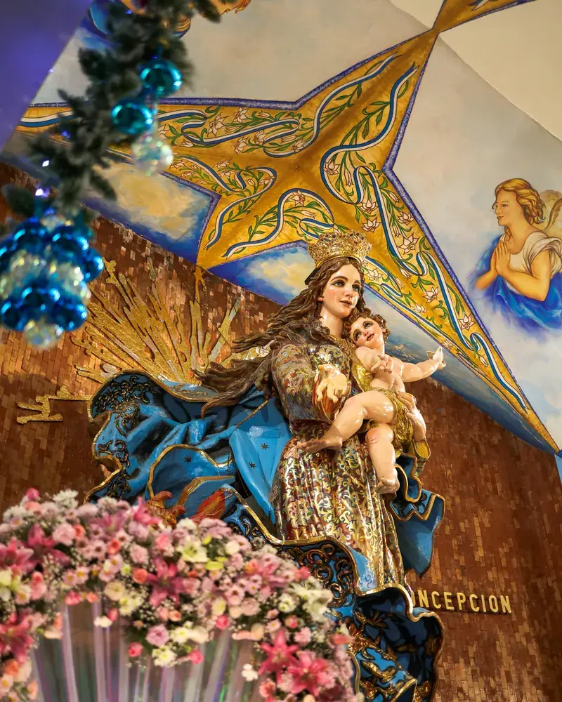 La imagen de la Inmaculada Concepción adornada con flores rosas y blancas a sus pies, vista desde abajo.