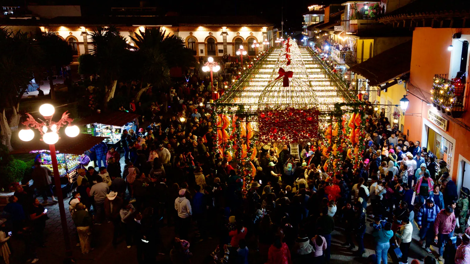 The Alley of Eternal Christmas at night seen from a drone: the crowded street leads to the tunnel of lights, with its arch of red ornaments, next to the plaza