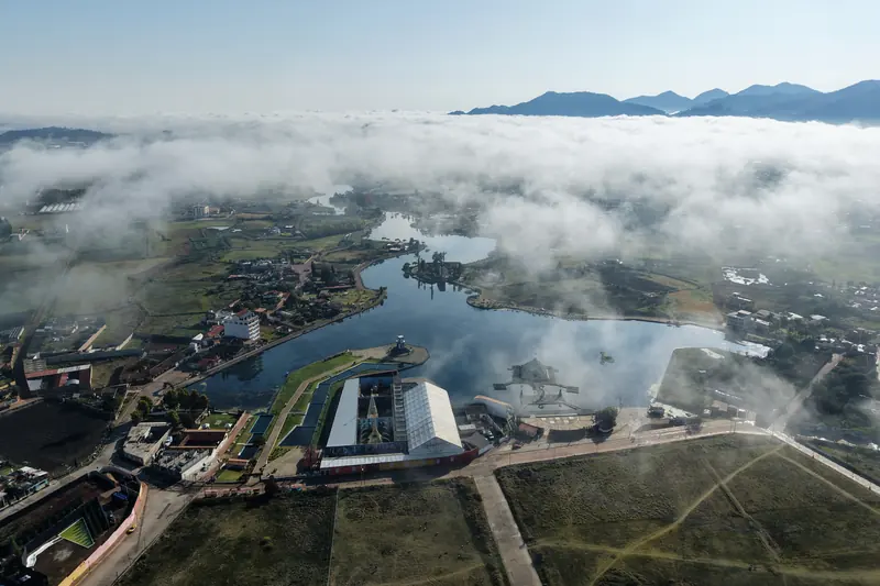 Vista aérea de la laguna de Chignahuapan en una mañana de niebla: un mar de nubes cubre el valle y deja ver el agua y el malecón, con la sierra asomando al fondo.