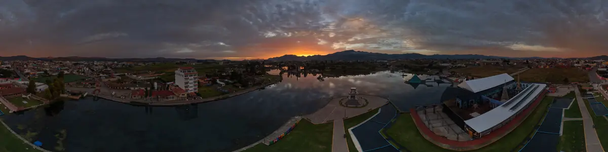 Panorámica aérea de la laguna de Chignahuapan al amanecer, con el sol saliendo detrás de la sierra y el pueblo alrededor del agua.