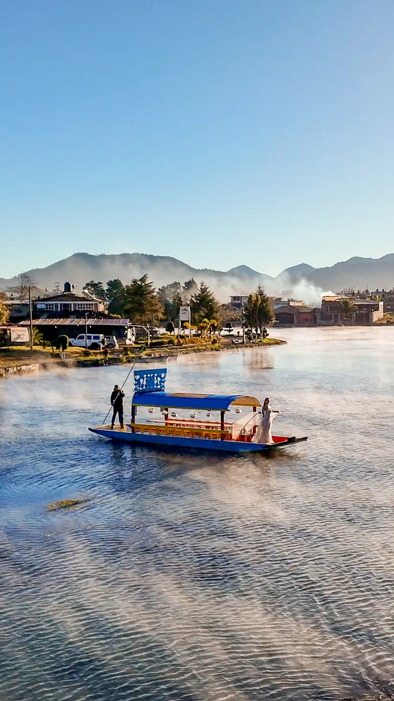 La laguna de Chignahuapan al amanecer: la niebla se levanta del agua mientras una lancha de colores cruza, con el malecón, el bosque y la sierra al fondo.