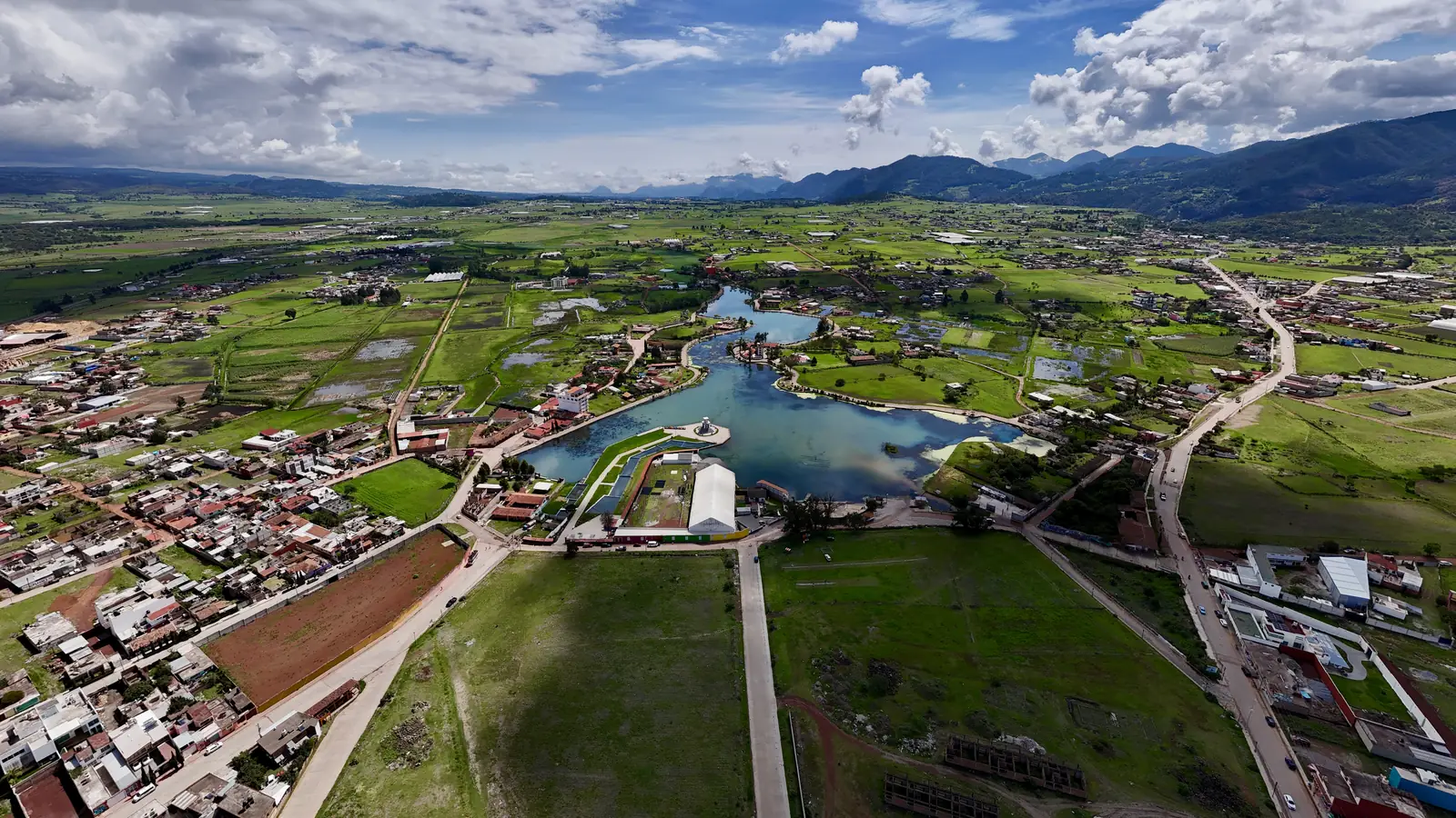 Chignahuapan from the air: the road entering the valley beside the lagoon, between green fields, with the mountains behind