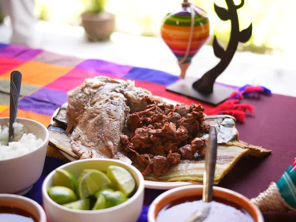 Barbacoa servida sobre pencas de maguey, con limones, arroz y salsas en una mesa de mantel de colores.