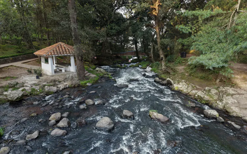El río Quetzalapan corre entre piedras junto a una palapa blanca con techo de teja, rodeado de árboles.