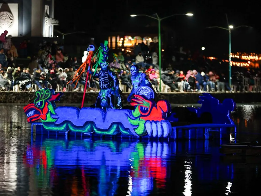 An illuminated raft with a colorful serpent and skull figures sails across the lagoon at night, in front of packed stands