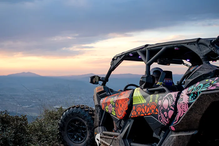 An off-road RZR decorated with colorful skulls stops on a hilltop at sunset, overlooking the valley and the mountains
