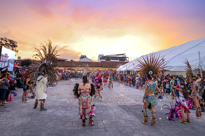 Dancers in feathered headdresses and colorful embroidered costumes perform at sunset, surrounded by visitors
