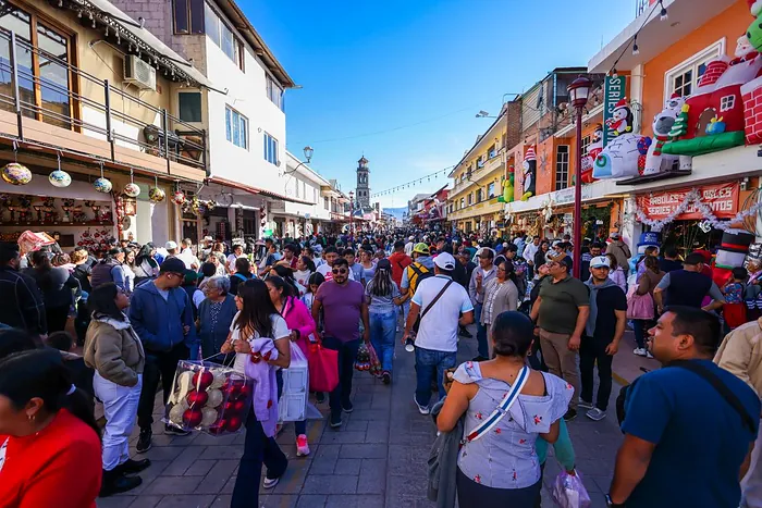 A downtown Chignahuapan street full of visitors on a sunny day, lined with shops selling Christmas decorations, with a church tower in the background