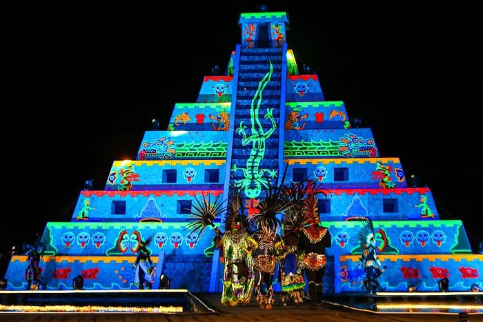 La pirámide del festival encendida en azul y verde, con danzantes de penacho al frente