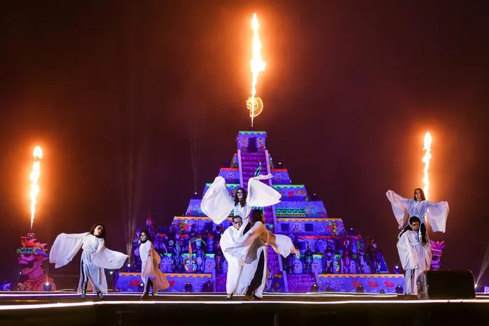 Dancers with white veils like wings in front of the pyramid lit in purple, with flaming torches above, during the Festival of Light and Life show