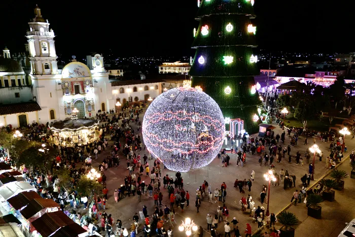 La esfera y el árbol monumentales encendidos frente a la Parroquia, con la plaza llena de gente de noche