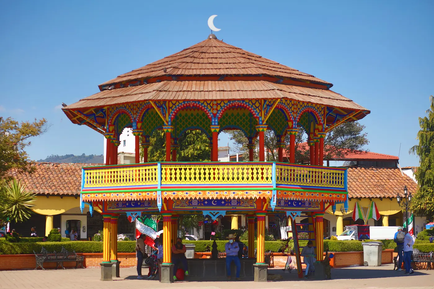 El kiosco de madera del centro de Chignahuapan visto desde la plaza: techo de teja, arcos de colores y barandal amarillo bajo el cielo azul.