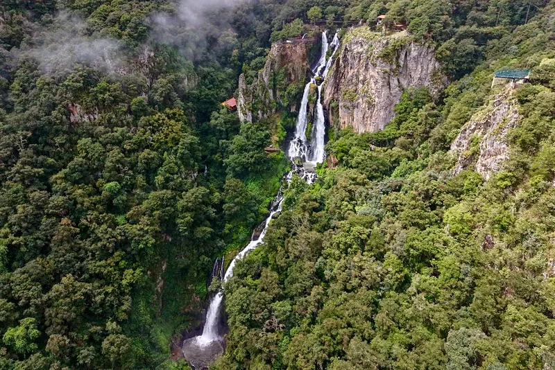 La cascada de Quetzalapan vista desde un costado y desde lo alto: el agua baja en diagonal junto a una pared de roca, entre el bosque y la neblina.