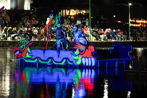 Una barca iluminada con calaveras y cabezas de serpiente de colores fosforescentes flota de noche en la laguna, reflejada en el agua, frente al público