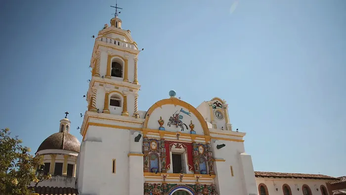 The facade of the Parish of Santiago Apóstol (St. James the Apostle) with its tower, dome and painted indigenous-Baroque reliefs, under a blue sky