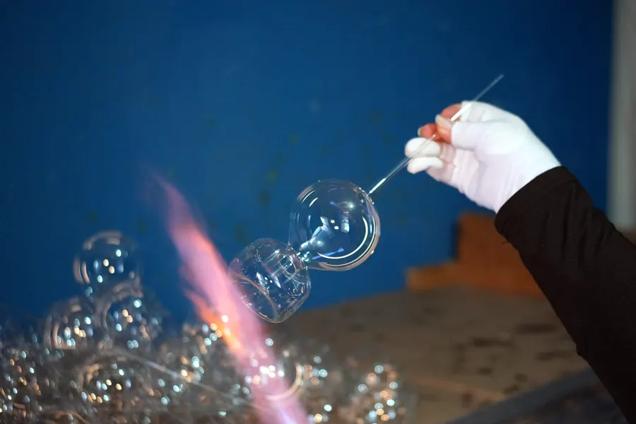 Una mano con guante blanco sostiene la varilla de vidrio con dos burbujas recién sopladas junto a la flama del soplete; abajo, un montón de esferas transparentes