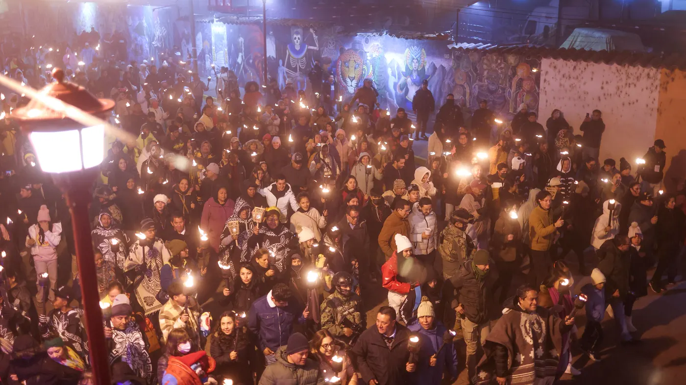 Una multitud abrigada camina de noche por la calle con cientos de antorchas encendidas rumbo a la laguna, junto a un farol