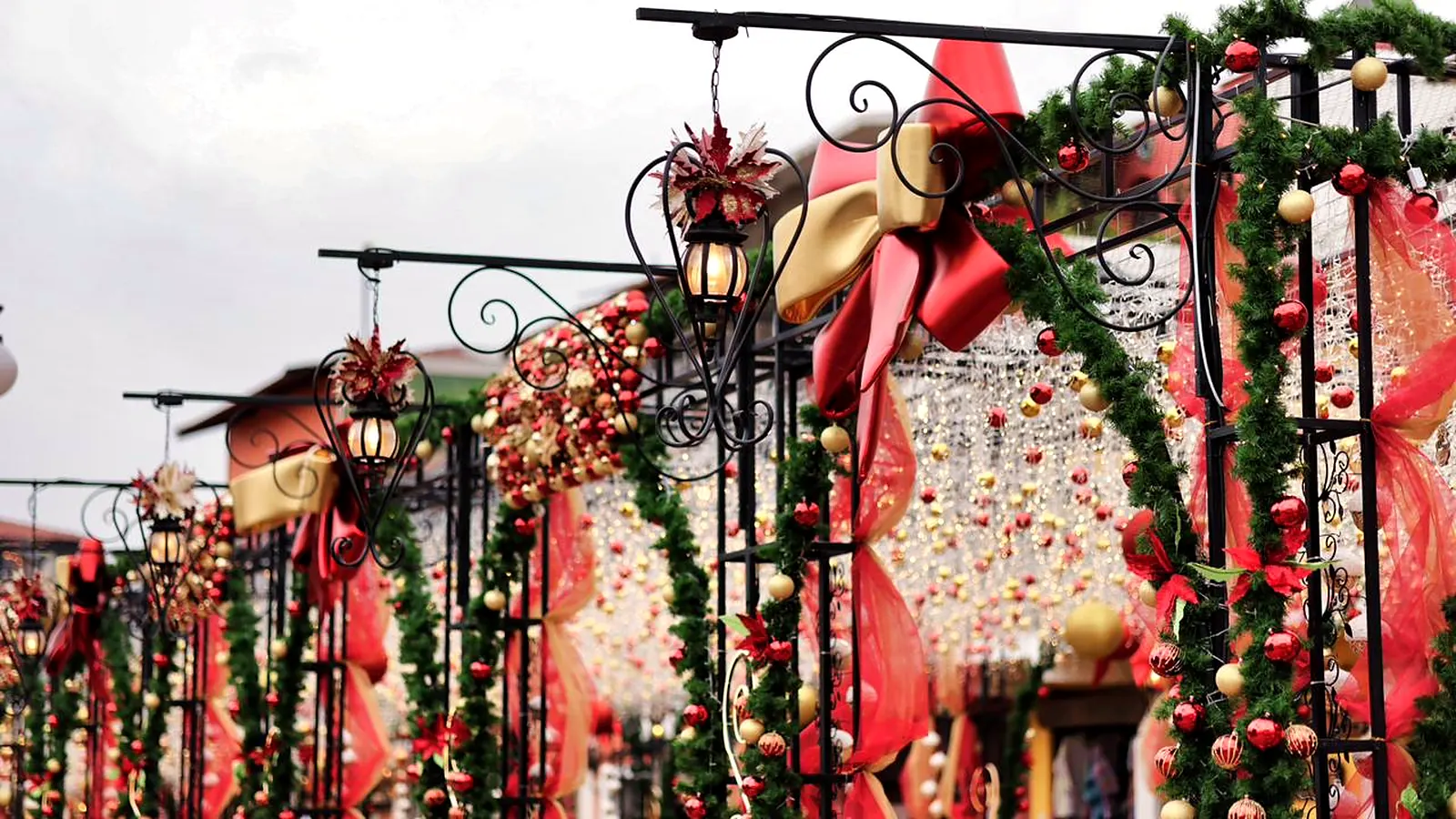 The Eternal Christmas Alley by day, seen from street level: wrought-iron arches with lanterns, green garlands, big red and gold bows and a curtain of ornaments hanging over the walkway
