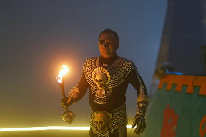 Un personaje de la puesta en escena, con el rostro pintado de calavera y un traje negro adornado con calaveras, avanza entre la bruma con una antorcha encendida