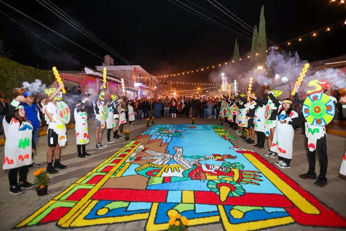 A colorful sawdust carpet with a pre-Hispanic figure covers the street at night, between rows of young people in painted white ponchos holding smoking incense burners, staffs and shields, during the Festival of Light and Life