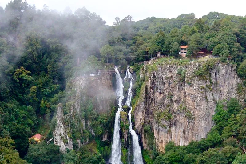 La parte alta de las Cascadas de Quetzalapan entre la niebla: el agua cae al centro por la pared de roca, rodeada de bosque