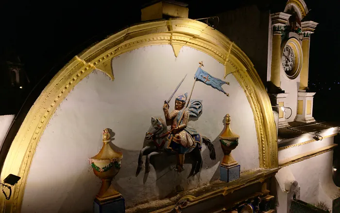 El remate de la fachada de la Parroquia iluminado de noche: Santiago a caballo con su estandarte, entre dos jarrones, bajo el arco dorado.