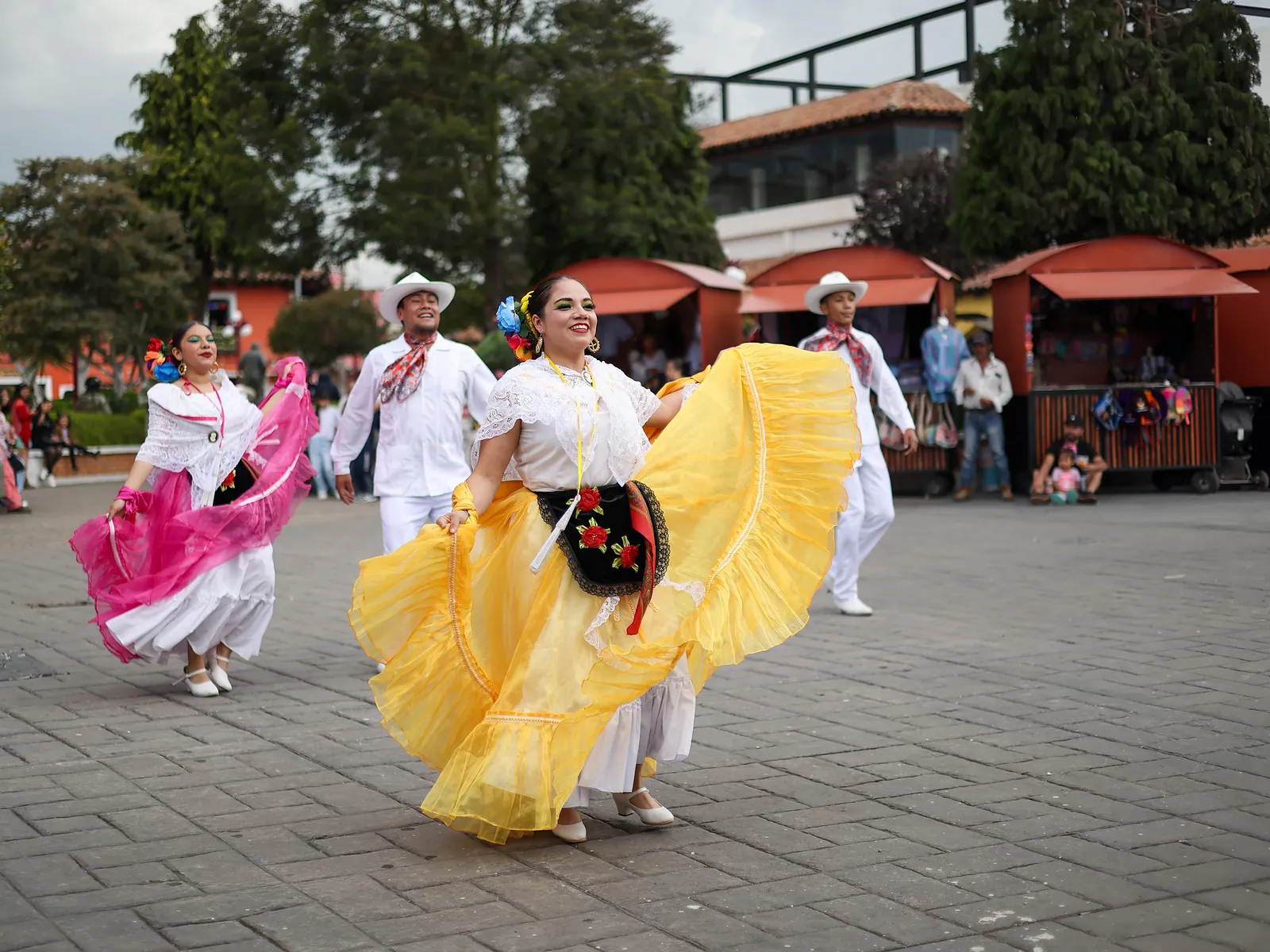 Bailarinas de folclor con faldas amarillas y rosas giran en la plaza de Chignahuapan durante una fiesta, entre puestos y árboles