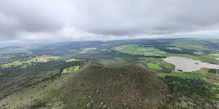 Un cerro de forma cónica cubierto de bosque y pasto, entre parcelas y un cuerpo de agua, bajo un cielo nublado.
