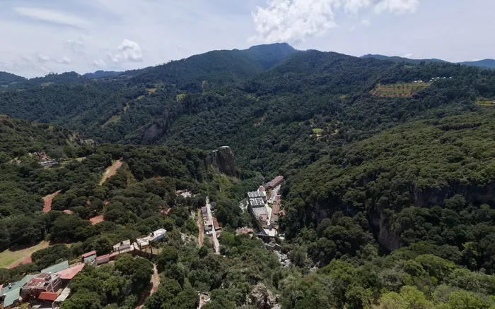 La barranca de las aguas termales desde el aire: un cañón cubierto de bosque con construcciones al fondo y la sierra detrás.