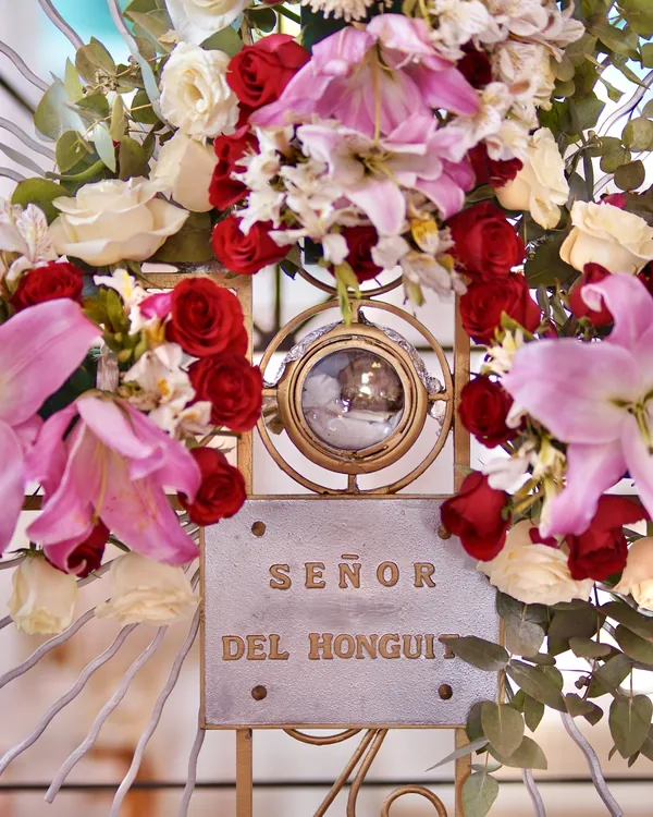 El relicario dorado del Señor del Honguito, con su placa, rodeado de rosas rojas, lirios rosas y flores blancas.