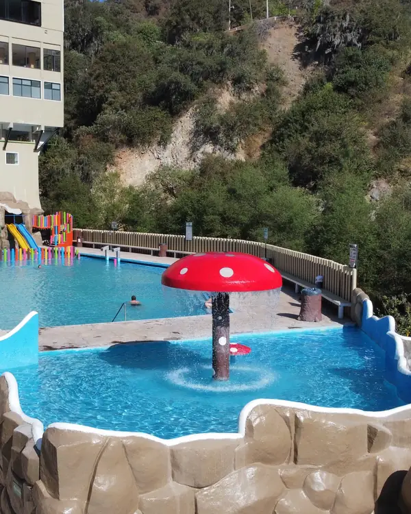 A shallow thermal paddling pool with a red mushroom-shaped fountain, in front of a pool with slides and the forested hill behind.