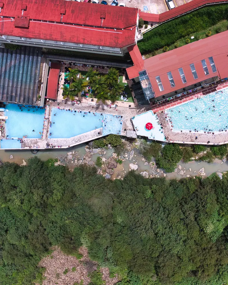The thermal pools seen from above, on the edge of the river that runs over stones along the bottom of the ravine, with the forest opposite.